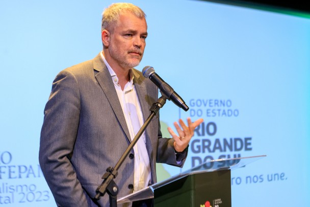 Alexandre Elmi está vestido de casaco cinza e camisa branca e gesticula com a mãe enquanto fala no microfone