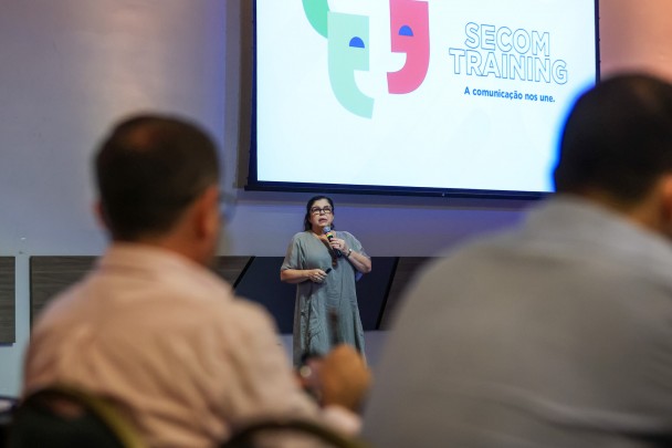Pessoas de costas observam a secretária Tânia falando no microfone