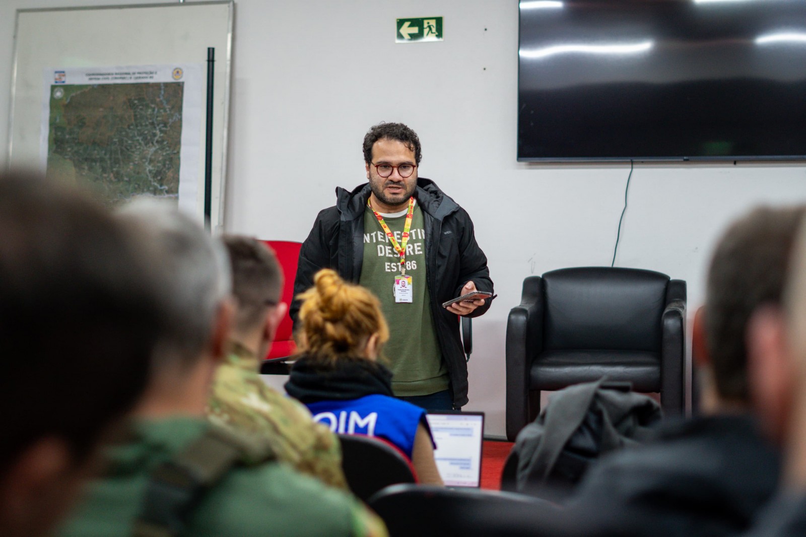 A foto mostra o repórter Thales Moreira de pé em um ambiente interno, em um centro de operações no Vale do Taquari. Ele é um homem de cabelo cacheado e óculos, vestindo uma jaqueta preta sobre uma camiseta verde oliva e segurando um celular na mão direita. Um crachá está pendurado em seu pescoço. À sua frente, algumas pessoas estão sentadas de costas, prestando atenção à sua fala ou apresentação. Ao fundo, uma lousa branca exibe um mapa, e um grande monitor de TV está pendurado na parede. No topo, uma placa verde indica uma saída de emergência.