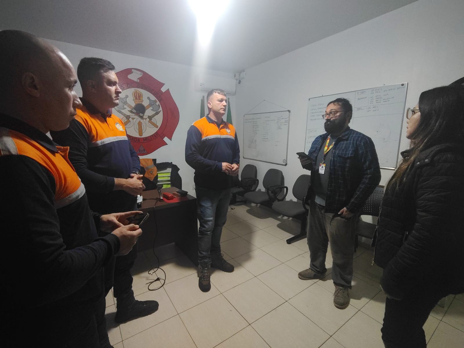 A foto exibe um grupo de cinco pessoas em uma sala de escritório com paredes brancas. O repórter Daniel Marcilio em pé, cercado pelas outras pessoas. Ele é um homem barbudo com óculos, camisa xadrez e casaco escuro segura um celular. Ao seu lado direito há uma mulher. Ao lado esquerdo, estão outros três homens. Eles vestem camisetas laranjas e azuis, semelhantes a uniformes. Ao fundo, há uma mesa com papéis e, na parede, um emblema circular e um quadro branco com anotações. A cena sugere uma reunião de trabalho.