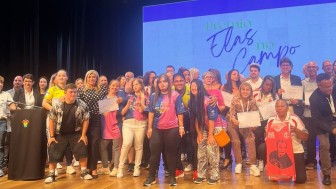 Esta &eacute; uma descri&ccedil;&atilde;o da imagem com foco em acessibilidade:

A fotografia colorida mostra um grande grupo de pessoas reunidas no palco de um teatro ou audit&oacute;rio para uma cerim&ocirc;nia de premia&ccedil;&atilde;o.

Detalhes da cena:

O Grupo: Cerca de 30 pessoas de diversas idades e perfis ocupam o centro do palco. Muitas delas sorriem e seguram certificados de papel branco. H&aacute; uma presen&ccedil;a not&aacute;vel de jovens com camisas esportivas coloridas (tons de rosa, azul e amarelo) e adultos em trajes casuais e formais.

Destaques: &Agrave; direita, uma mulher negra sentada &agrave; frente do grupo segura uma camiseta regata vermelha com a estampa de um rosto e a frase "RAUL VIVE". &Agrave; esquerda, uma mulher loira com blusa preta de bolinhas brancas abra&ccedil;a uma das jovens premiadas.

O Cen&aacute;rio: Ao fundo, um grande tel&atilde;o exibe um fundo azul claro com a frase escrita em letras cursivas e elegantes: "Pr&ecirc;mio Elas no Campo". O palco tem piso de madeira clara e est&aacute; iluminado por diversos refletores no teto.

Elementos Laterais: &Agrave; extrema esquerda, v&ecirc;-se um p&uacute;lpito de madeira escura com um bras&atilde;o colorido e um microfone, indicando o local dos discursos.

Ilumina&ccedil;&atilde;o e Clima: A luz &eacute; vibrante e festiva, capturando um momento de celebra&ccedil;&atilde;o coletiva e reconhecimento. A foto possui um &acirc;ngulo frontal que abrange quase toda a largura do grupo premiado.